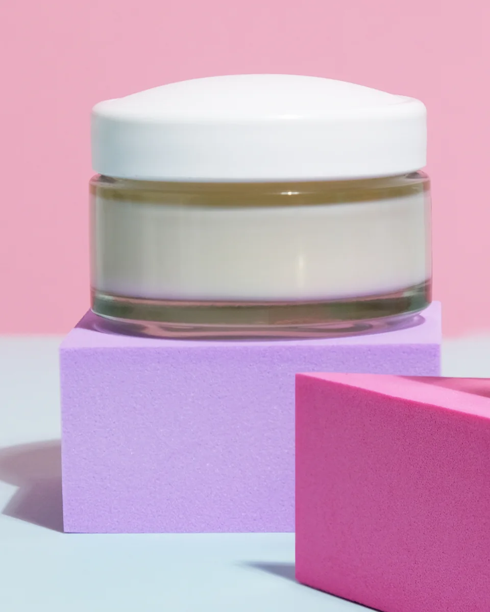 A glass jar of cream with a white lid on a lilac block