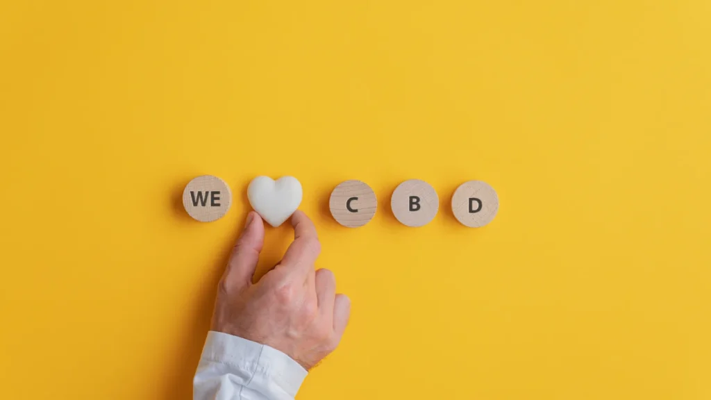 Cannabis content marketing: a hand places a white heart between wooden tiles that read WE and CBD, on a yellow background