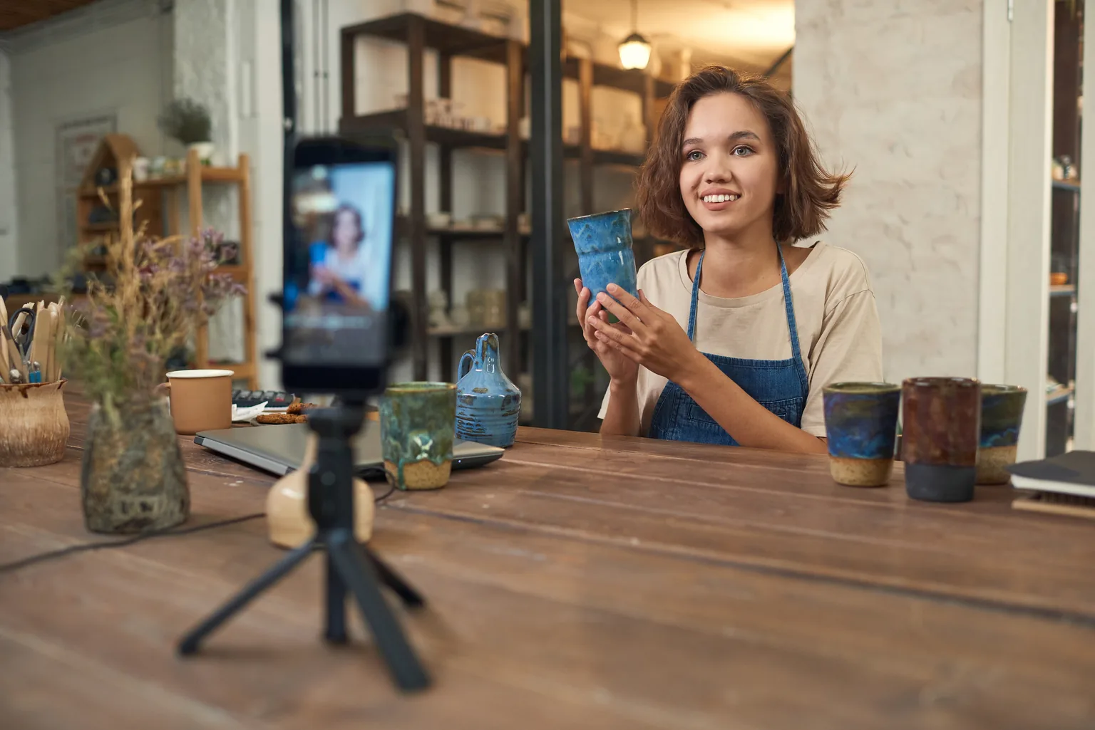 A small business owner in an apron films a live video of handmade cups with a phone on a tripod
